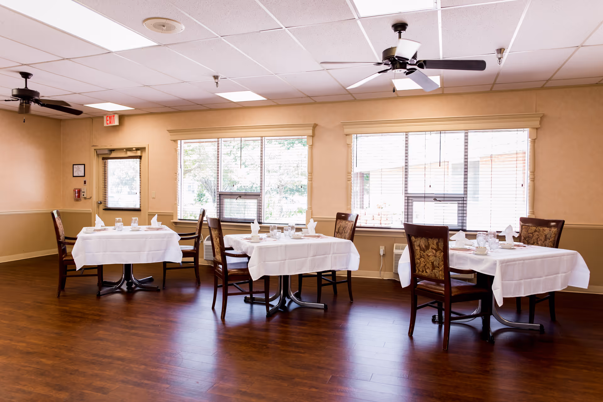 A dining room with three tables covered in white tablecloths, each set with glasses, napkins, and plates. The room has large windows with blinds, wooden floors, beige walls, and ceiling fans.
