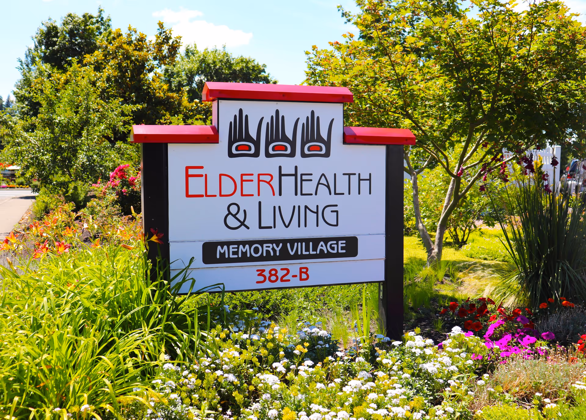 Outdoor sign for ElderHealth & Living Memory Village with red and black text and hand symbols, surrounded by green plants, colorful flowers, and trees under a blue sky.
