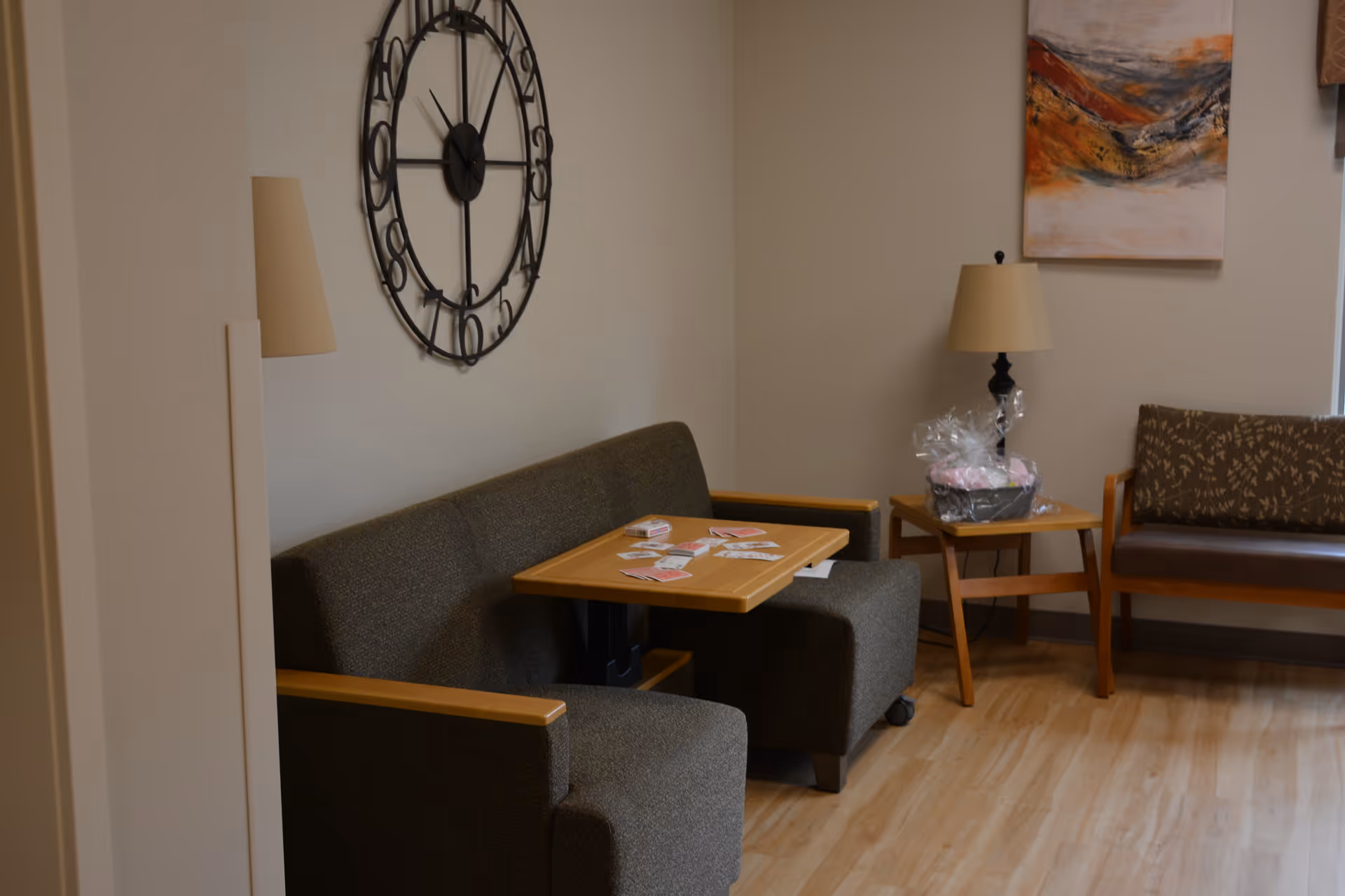A cozy seating area in a senior living facility with a dark green upholstered sofa and chair around a wooden table with playing cards on it. There is a large decorative wall clock above the sofa, a side table with a lamp and a wrapped gift basket, and another bench with patterned cushions against the wall. The floor is light wood, and there is a colorful abstract painting on the wall.