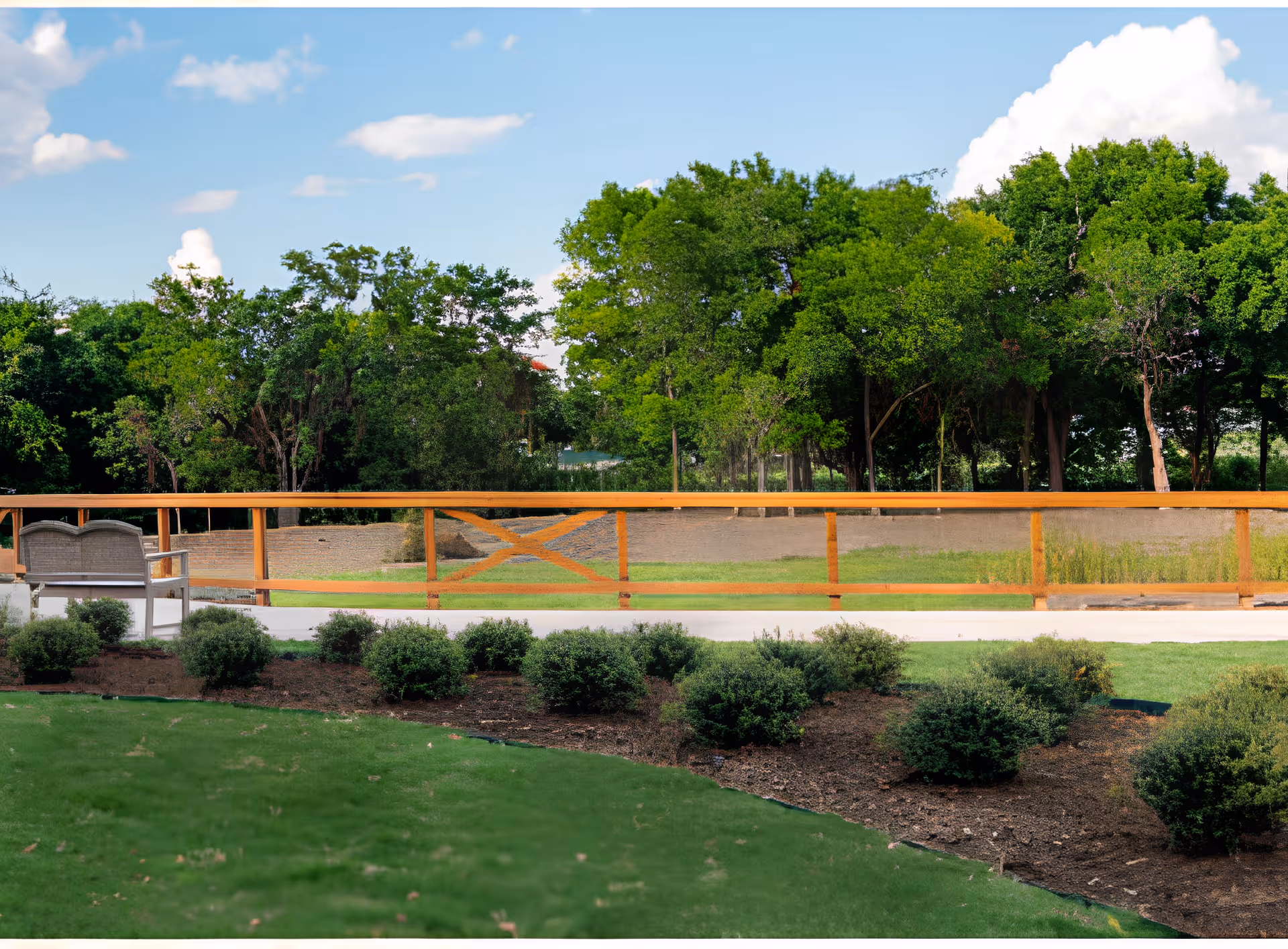Landscaped outdoor courtyard with a wooden railing, bench seating, trimmed shrubs and trees in the background.