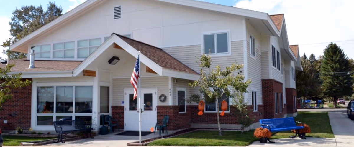 Exterior view of Meadowlark Assisted Living facility showing a two-story building with white siding and red brick accents. There is a small covered entrance with an American flag on a pole in front. Benches and small trees with orange decorations are visible on the lawn near the entrance. A driveway and parked car are seen on the right side.
