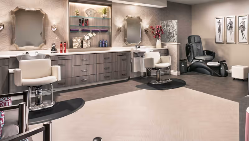 Interior of a salon area with two white salon chairs in front of sinks and mirrors mounted on a wall with decorative wallpaper. Shelves with hair products and decorative items are visible above the cabinets. To the right, there is a black pedicure spa chair and three framed fashion sketches on the wall.