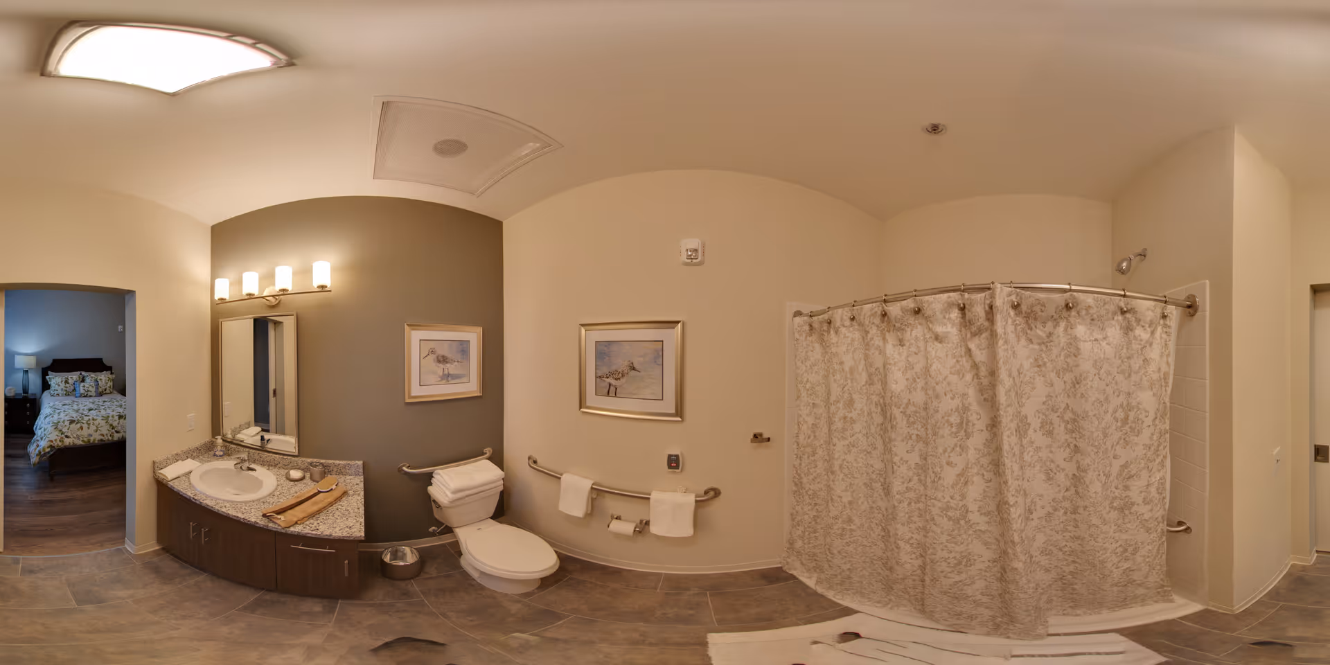 A spacious bathroom with a granite countertop sink, a large mirror with four lights above it, a toilet with grab bars on the wall, and a shower with a floral-patterned curtain. The bathroom has beige and gray walls, tile flooring, and a doorway leading to a bedroom with a bed and nightstand visible.