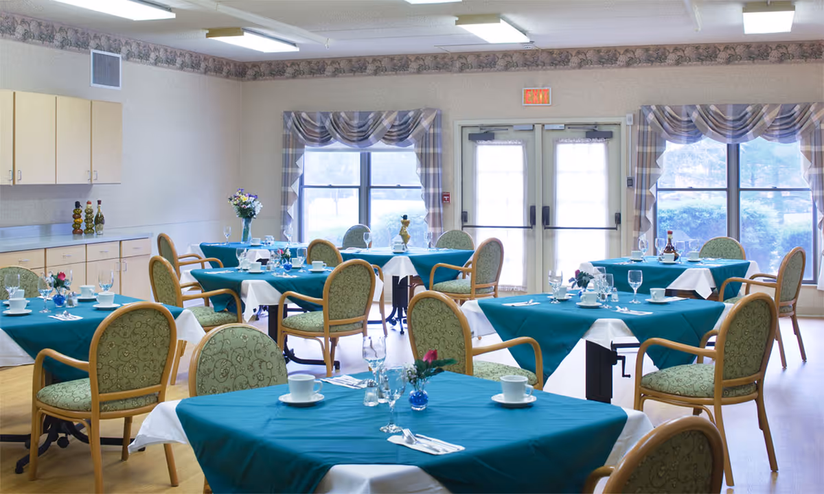 Dining room with multiple round and square tables covered in teal tablecloths, place settings and chairs in a bright room with windows and double doors.