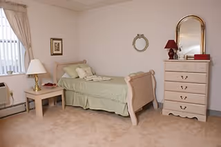 A simple single-occupancy bedroom with a bed, nightstand and lamp, dresser with mirror, and a curtained window.