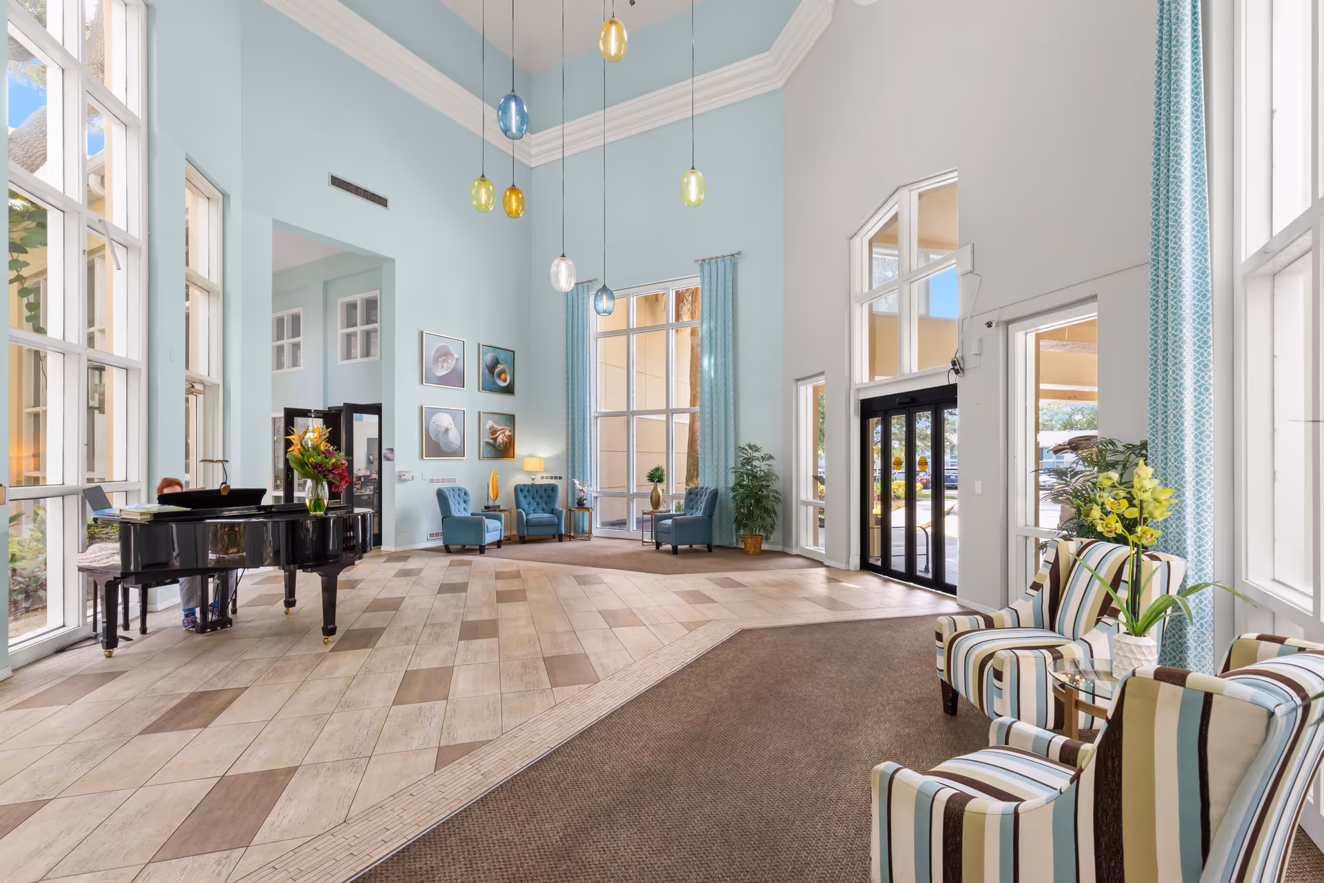 Bright and spacious senior living facility lobby with high ceilings, large windows, and a mix of tiled and carpeted flooring. The room features a black grand piano with a person playing it, several blue armchairs arranged near a window with light blue curtains, striped armchairs with a glass side table and a flower vase, hanging colorful pendant lights, and framed artwork on the wall.
