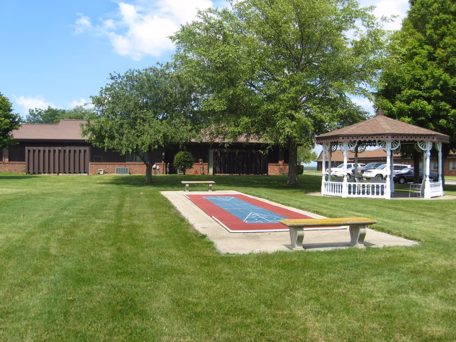 A well-maintained outdoor area at Vonderlieth Living Center featuring a shuffleboard court with benches on either side, a white gazebo with decorative trim, several trees providing shade, a grassy lawn, and a brick building in the background under a partly cloudy blue sky.