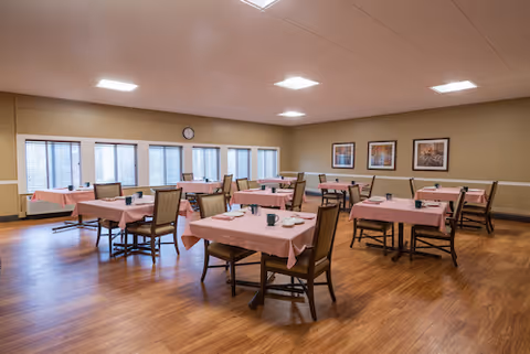 A spacious dining room with several tables covered in pink tablecloths, each set with plates, cups, and utensils. The room has wooden flooring, beige walls, and multiple windows letting in natural light. There are framed pictures on the wall and a clock above the windows.