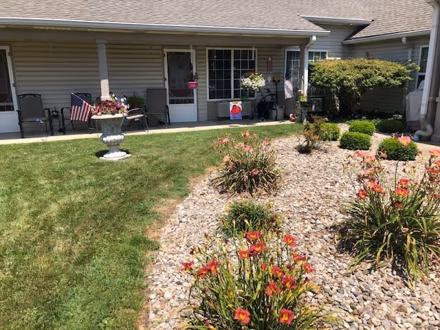A garden area with green grass and a rock bed containing flowering plants in front of a single-story building with a covered porch. The porch has chairs, potted plants, and an American flag. The building has beige siding and several windows and doors.