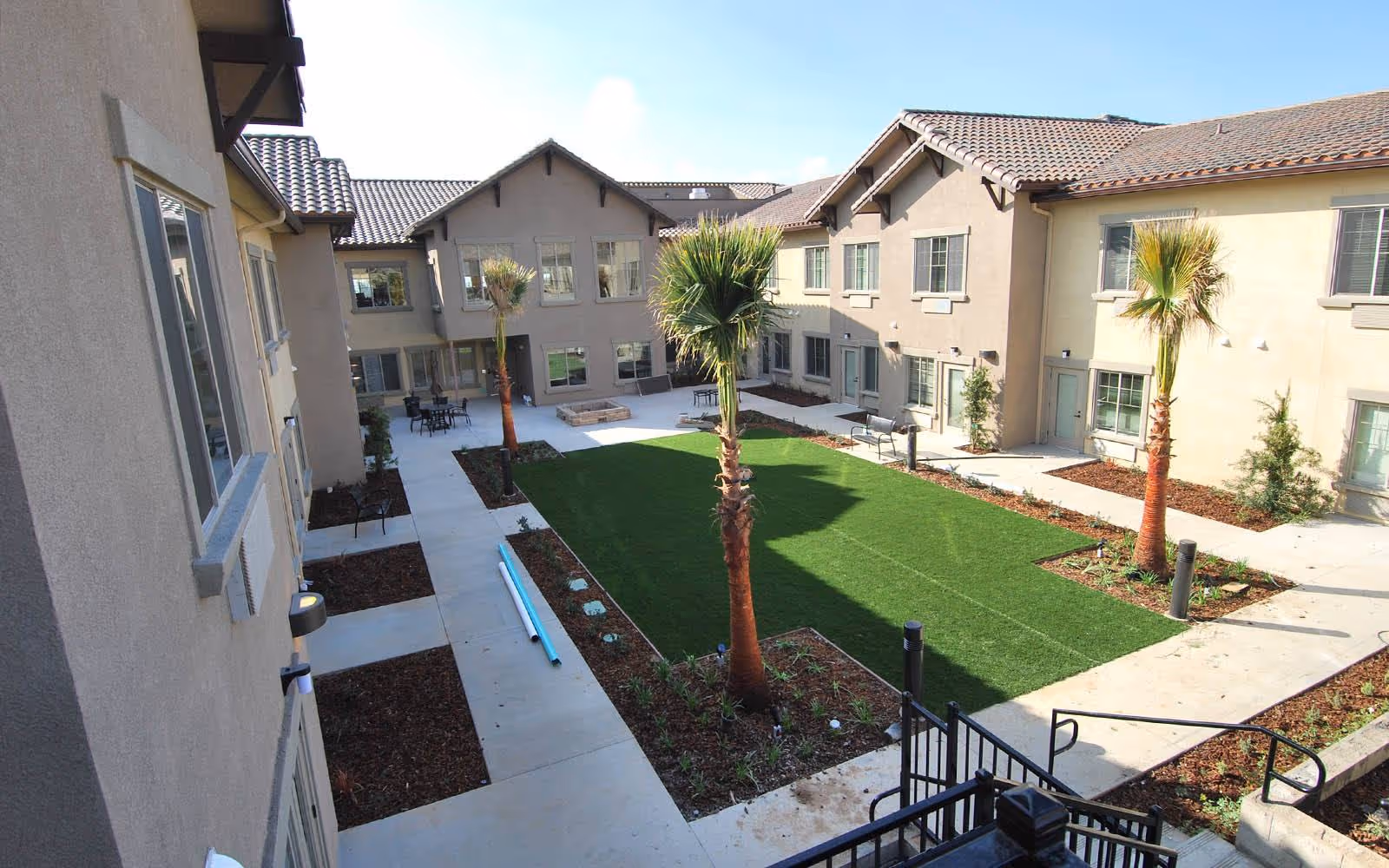 Outdoor courtyard area of a senior living facility with a green lawn, palm trees, benches, and surrounding two-story beige buildings with tiled roofs under a clear blue sky.