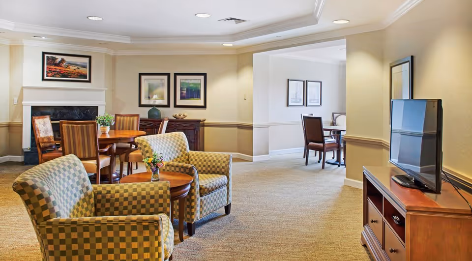 Communal lounge with patterned armchairs, round wooden tables, dining chairs, a TV on a wooden cabinet, and a fireplace.