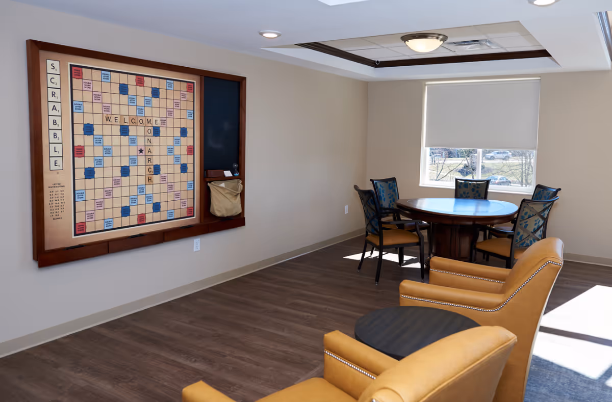 Bright common room with a large wall-mounted Scrabble board, round table and chairs, and two upholstered armchairs near a window.