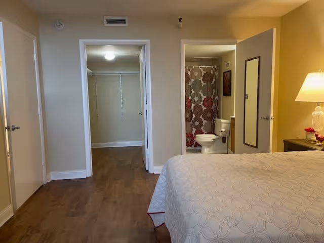 View of a bedroom with a bed covered in a white patterned bedspread in the foreground. To the right, there is a nightstand with a lamp and a small decorative item. In the background, there are two open doorways: one leading to a walk-in closet with shelves and hanging rods, and the other leading to a bathroom with a toilet and a shower curtain with a floral pattern.