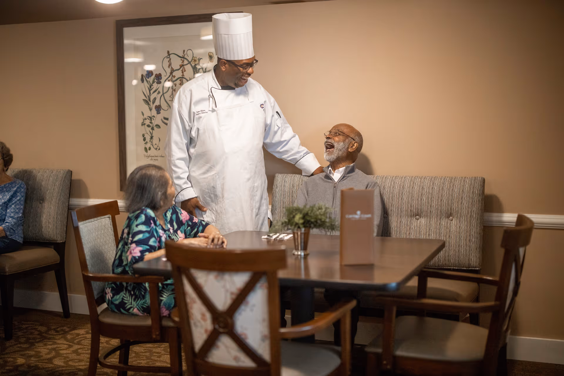 A chef in a white uniform and tall hat is smiling and talking to an elderly man seated at a dining table in a senior living facility. An elderly woman sits nearby, also at the table. The room has beige walls, framed artwork, and cushioned chairs.