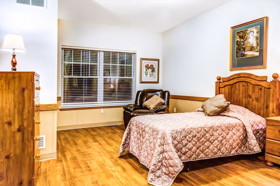 A furnished bedroom with a wooden bed, dresser, armchair, lamp, window with blinds, and framed artwork.