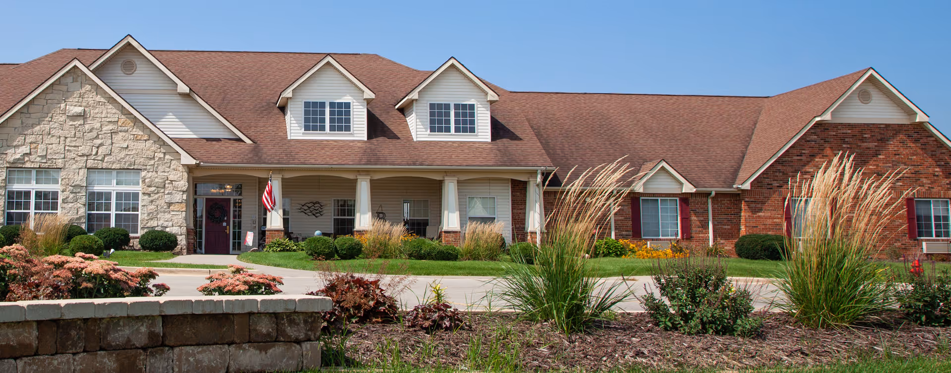Single-story red brick and stone building with a covered front porch, dormer windows, driveway, and landscaped shrubs and ornamental grasses.