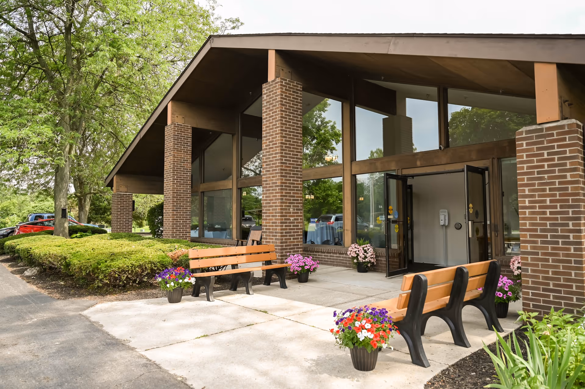 Front entrance of a brick senior care center with benches, potted flowers, large glass windows, and surrounding landscaping.
