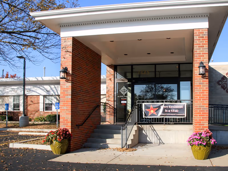 Entrance of a brick building with a covered porch supported by two brick columns. There are stairs leading up to glass doors with a banner that reads 'Every Resident is a STAR' and two large flower pots with pink and red flowers on either side of the stairs. The building appears to be part of a retirement community.