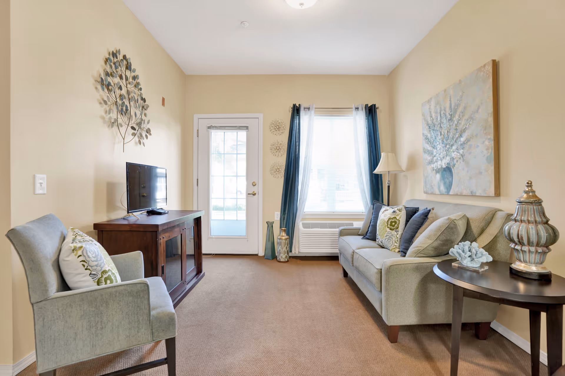 A cozy living room with beige walls and carpet. There is a light green upholstered armchair with a decorative pillow on the left, and a matching sofa with multiple pillows on the right. A wooden TV stand with a flat-screen TV is against the left wall. A window with blue and white curtains and a door with glass panels are at the far end of the room. Decorative items include a metal wall art piece above the TV, a large floral painting above the sofa, a floor lamp, and two vases on the floor near the window. A round side table with a decorative jar and coral piece is next to the sofa.