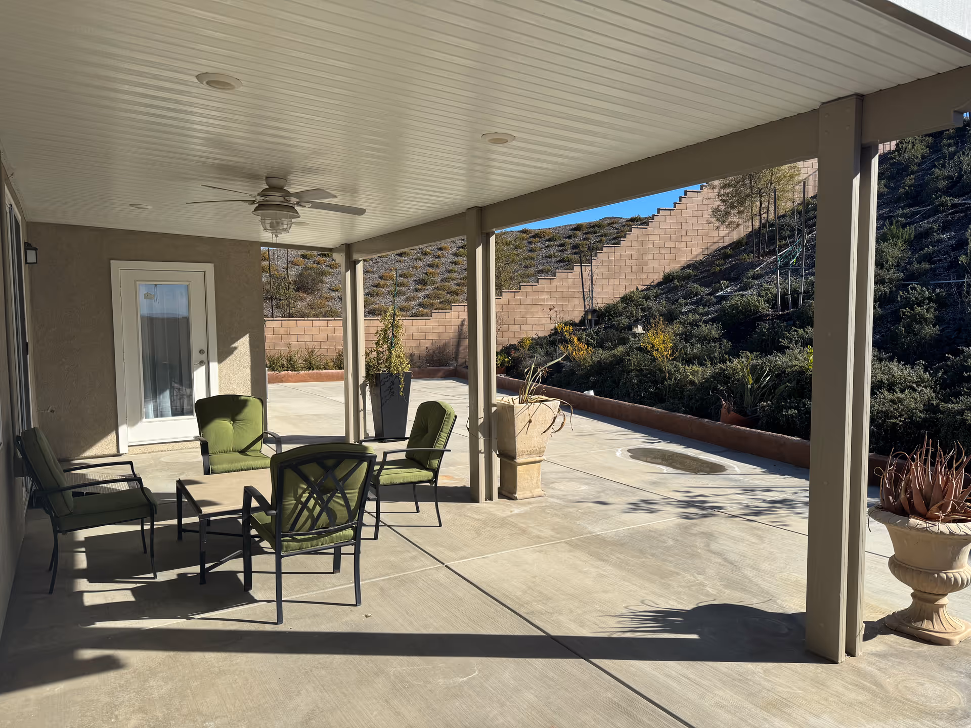 Covered patio with green-cushioned outdoor chairs, ceiling fan, and potted plants overlooking a sloped hillside and retaining wall.
