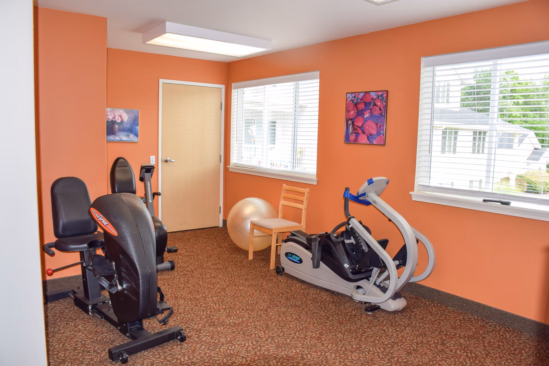 A small exercise room with orange walls and carpeted floor featuring two recumbent exercise bikes, a wooden chair, and a large exercise ball. Two windows with white blinds let in natural light, and there are two colorful paintings on the walls.