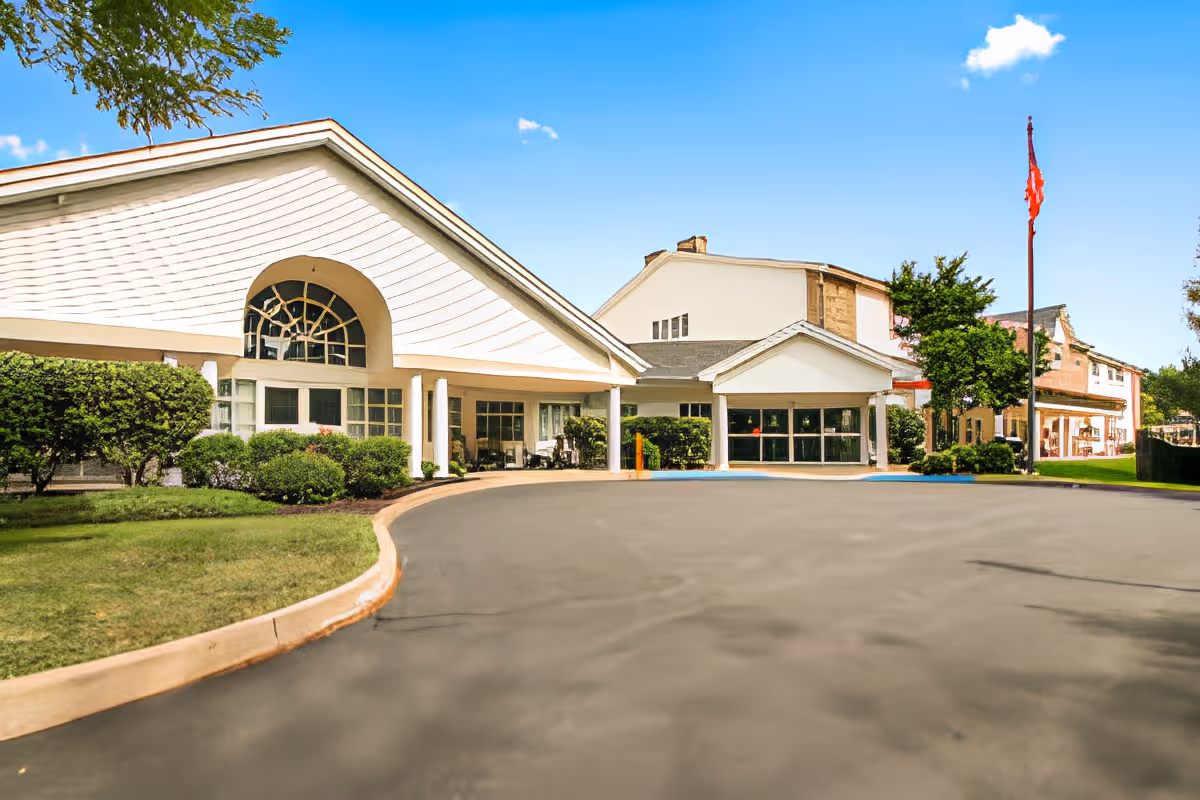 Exterior view of a senior living facility building with white walls, large windows, and a covered entrance. There are neatly trimmed bushes and a flagpole with a red flag on the right side. The sky is clear and blue.
