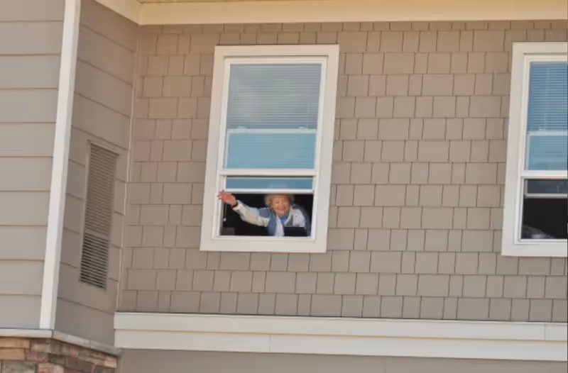 A person leaning out of a second-floor window of a beige shingled building, waving.