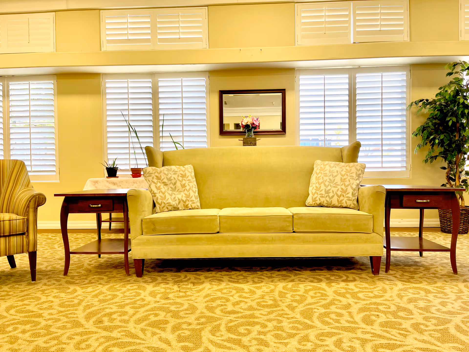 A cozy living room area with a light green sofa adorned with two patterned cushions, flanked by two wooden side tables. Behind the sofa are windows with white plantation shutters, a mirror on the wall, and a small vase with flowers. To the left is a striped armchair, and to the right is a tall potted plant. The floor is covered with a patterned beige carpet.