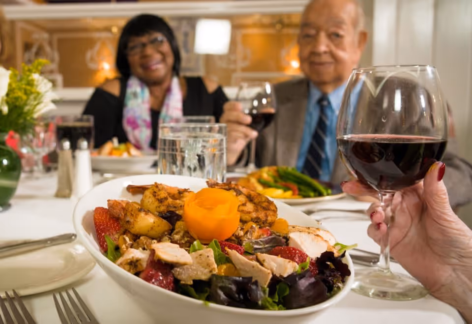 Two elderly people sitting at a dining table enjoying a meal together. In the foreground, there is a close-up of a salad with grilled chicken, strawberries, and a carved vegetable garnish, along with a hand holding a glass of red wine. The background shows the elderly man and woman smiling and holding glasses of wine.