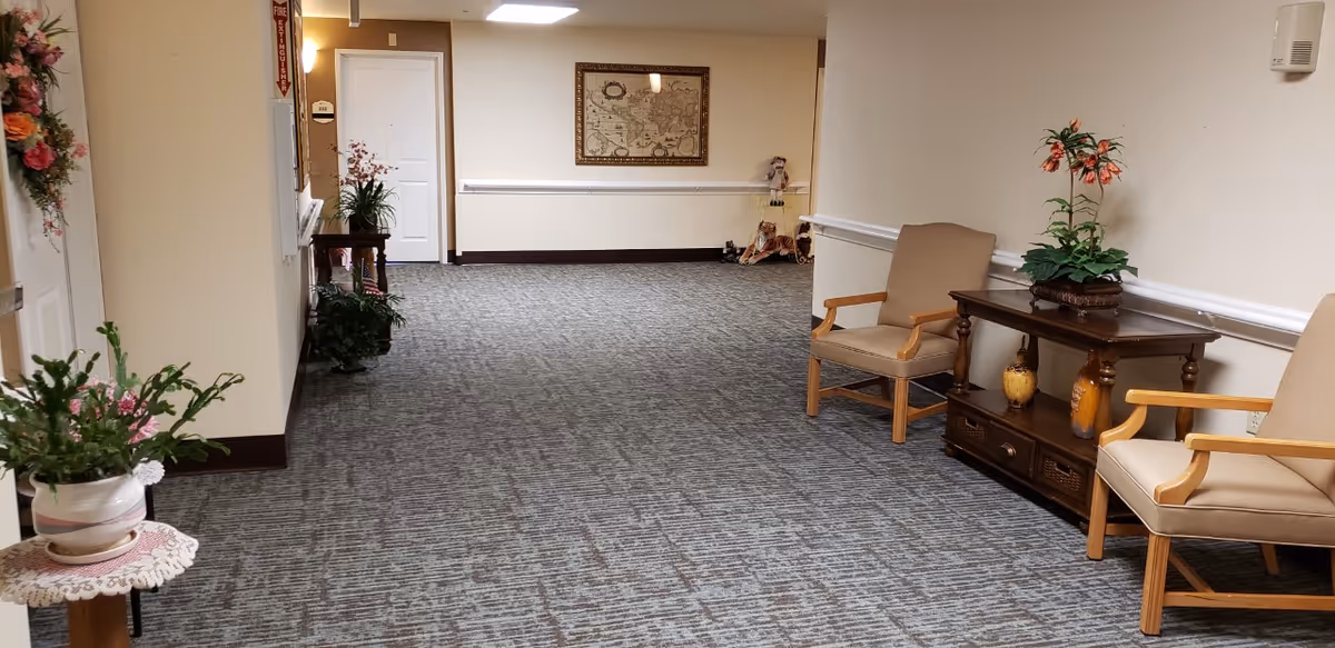 A carpeted hallway in a senior living facility with beige walls and white trim. There are two beige armchairs with wooden arms on the right side, separated by a wooden table holding a potted plant and decorative items. On the left side, there are several potted plants on stands and a fire extinguisher sign on the wall. At the end of the hallway, a framed map hangs on the wall above a small shelf with decorative items.