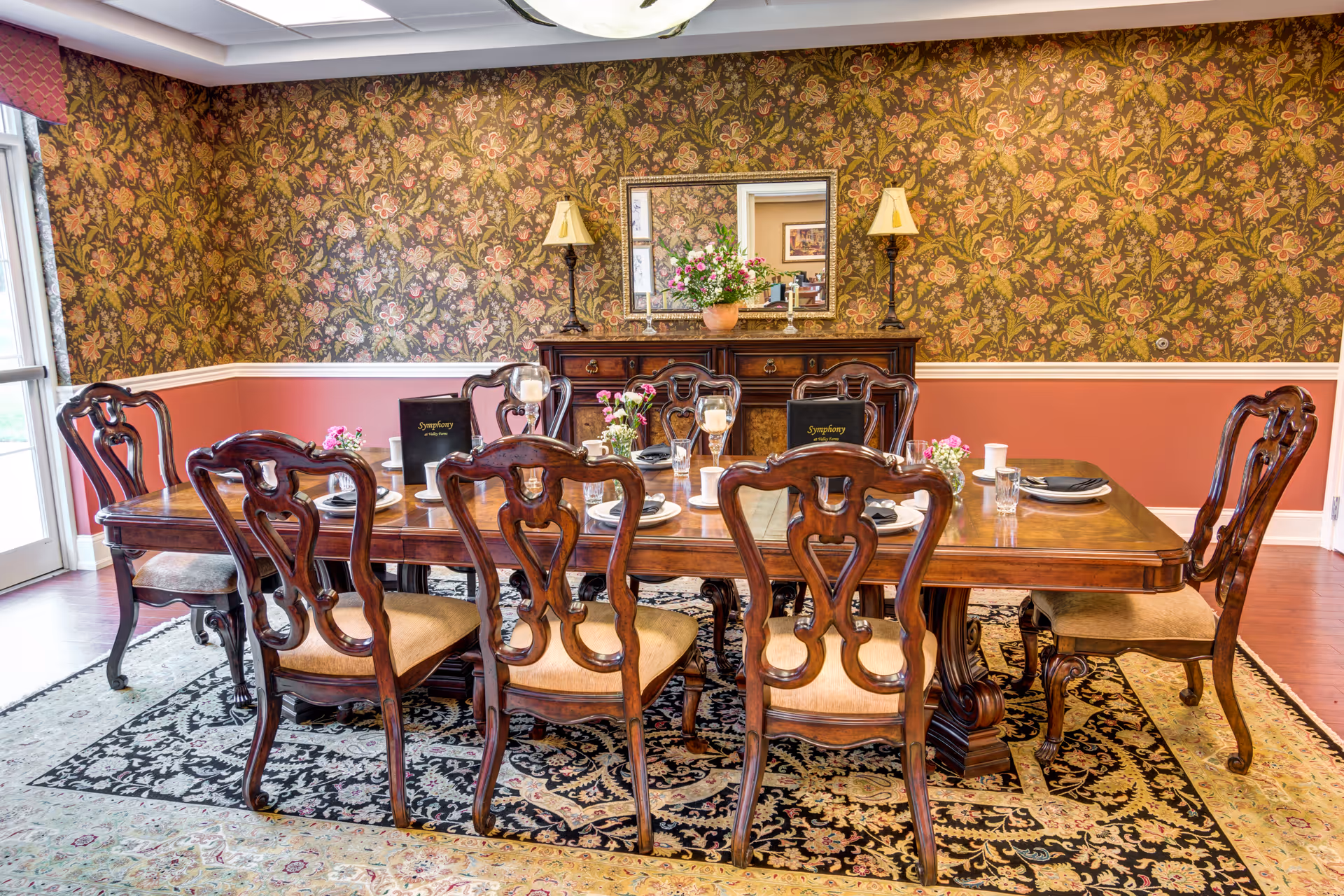A formal dining room with a large wooden dining table surrounded by eight ornate wooden chairs with cushioned seats. The table is set with plates, glasses, napkins, and small floral arrangements. Behind the table is a wooden sideboard with two lamps and a large mirror above it. The walls have floral wallpaper on the upper half and a solid reddish color on the lower half, with a chair rail separating them. A patterned area rug covers the floor beneath the table and chairs.