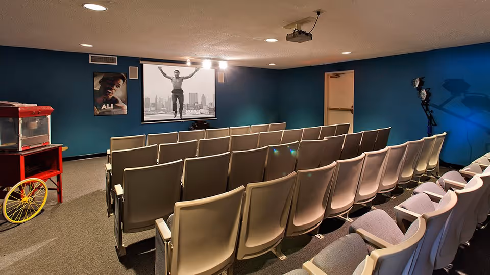 A small movie theater room with rows of beige seats facing a screen displaying a black and white image of a man jumping with a city skyline in the background. The walls are painted blue, and there is a popcorn machine on a red cart to the left. A poster of Muhammad Ali is on the left wall, and stage lights are positioned on the right side of the room.