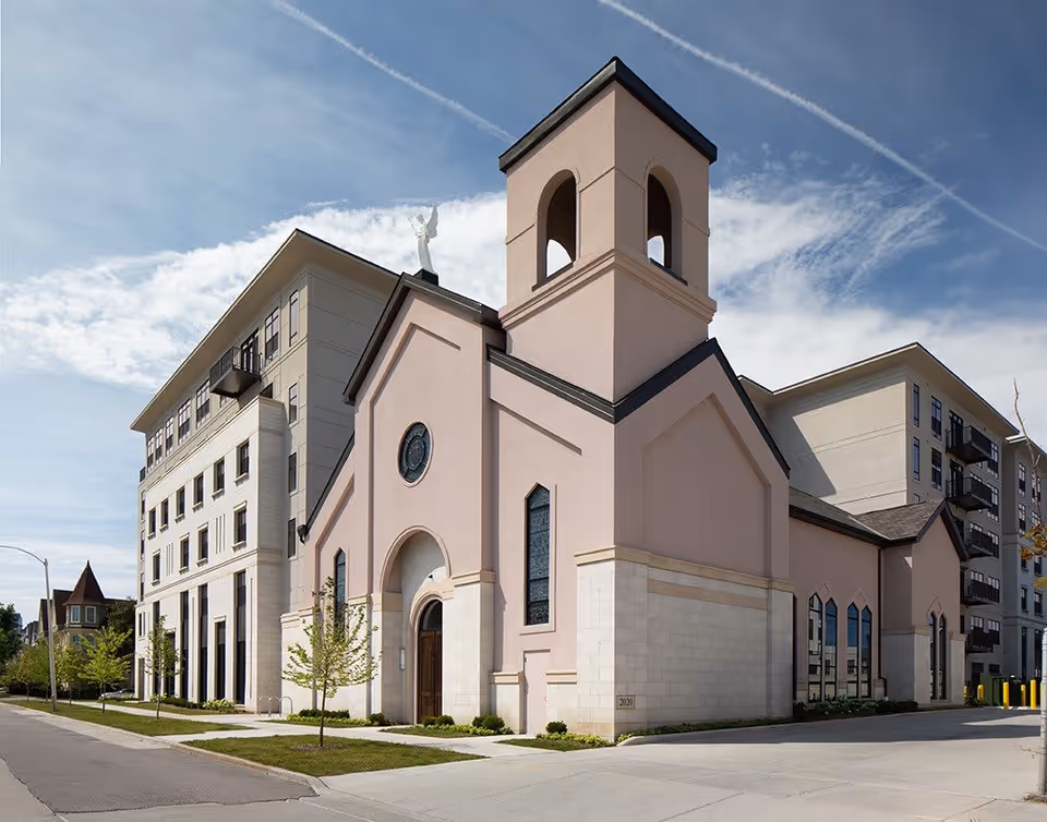 Exterior view of a pale pink, church-like building attached to a multi-story senior living complex under a blue sky.