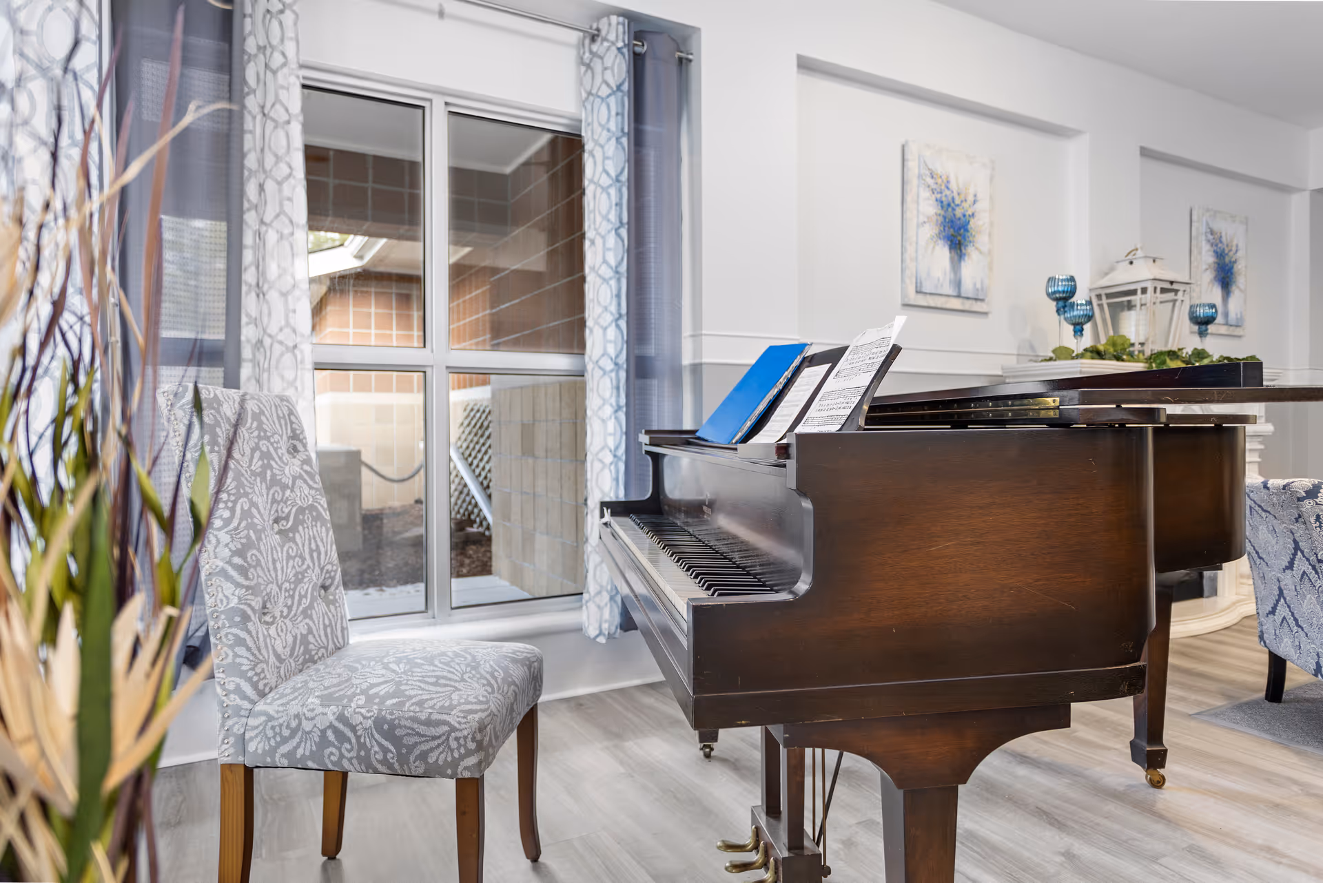 Interior view of a room featuring a dark wooden grand piano with sheet music on the stand, a patterned upholstered chair, large windows with patterned curtains, and decorative wall art and ornaments in the background.