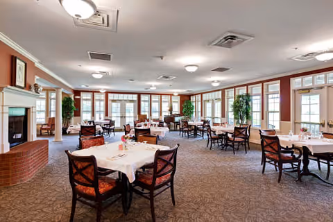 A spacious dining room with multiple tables covered in white tablecloths and set with small flower arrangements. The room features large windows along the walls allowing natural light to fill the space. There are several chairs around each table, patterned carpet flooring, and a brick fireplace on the left side. Potted plants are placed near the windows, and ceiling lights provide additional illumination.