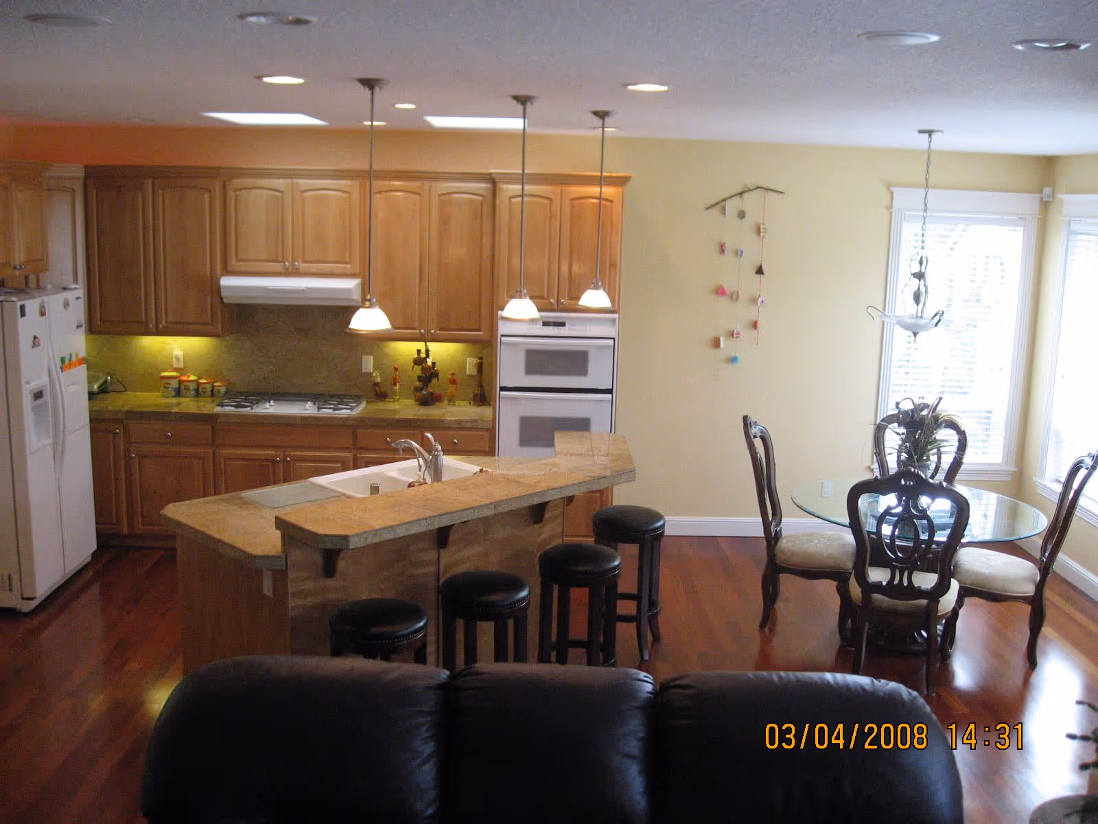 Interior view of a kitchen and dining area with wooden cabinets, a kitchen island with a sink and four black bar stools, a white refrigerator, a built-in oven, and a round glass dining table with four chairs near windows with blinds. The floor is wooden and there is a black leather couch partially visible in the foreground.