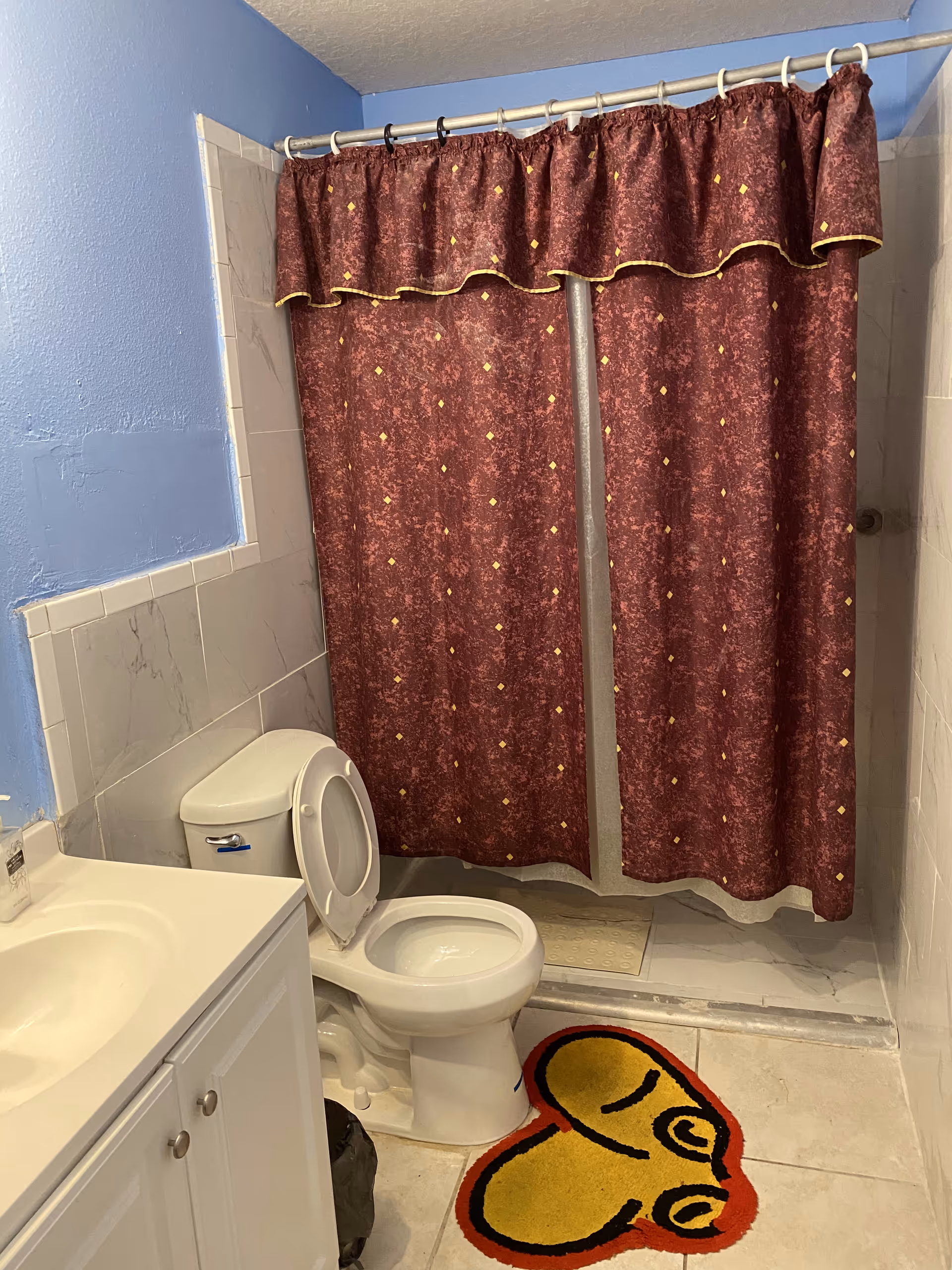 A bathroom with a white toilet with the lid open, a white vanity with a sink, a shower area with a maroon shower curtain featuring a subtle pattern and a valance, light blue painted walls, and a colorful yellow and red heart-shaped rug on the floor.