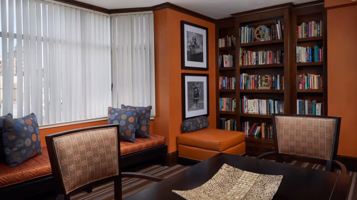 A cozy interior room with a large window covered by vertical blinds, an orange cushioned bench with patterned pillows, two framed black and white dog photographs on an orange wall, a wooden bookshelf filled with books, and a dark wooden table with two upholstered chairs and a decorative tray on top.
