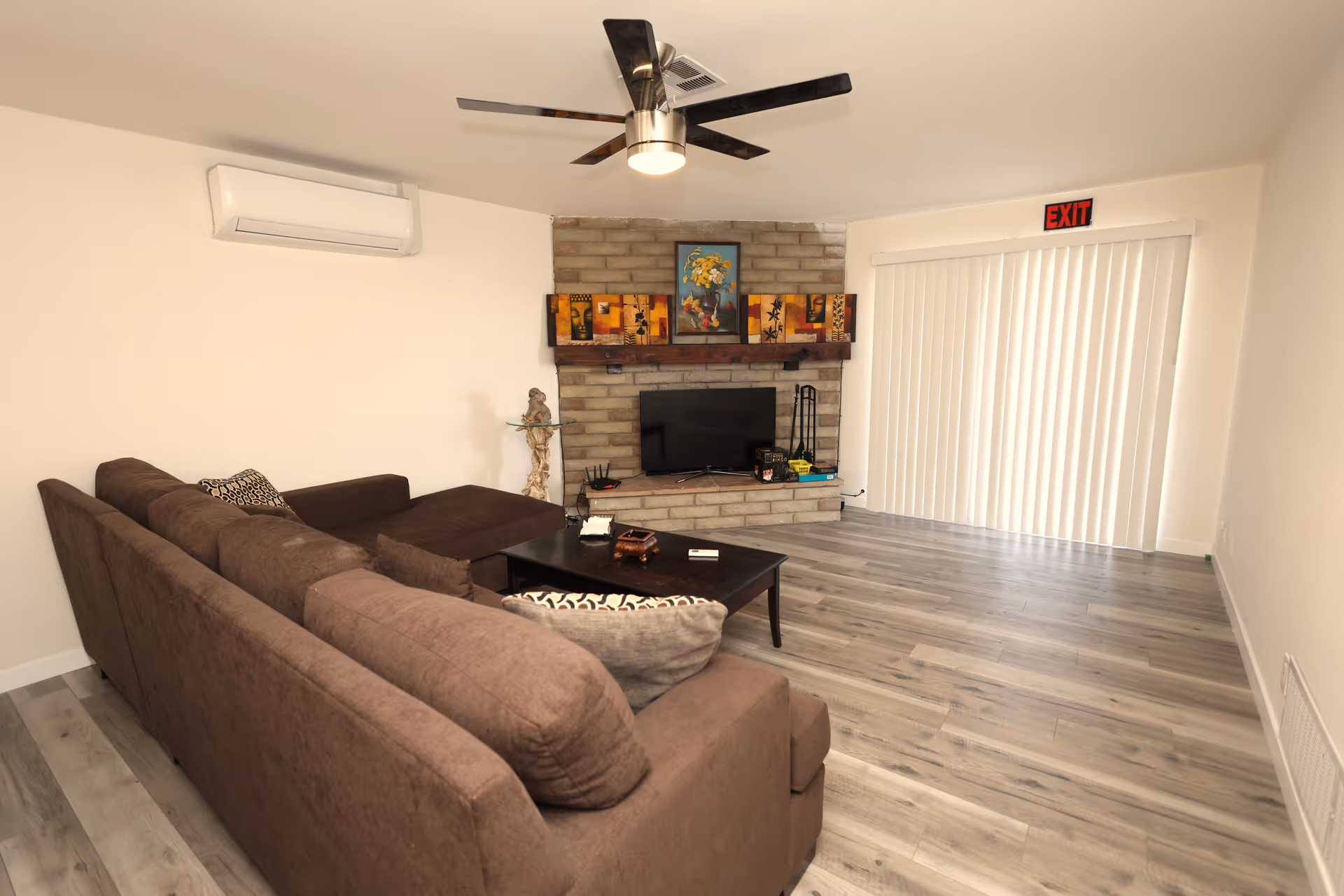 Living room with a brown sectional sofa, coffee table, TV on a brick fireplace, ceiling fan, and sliding vertical blinds.