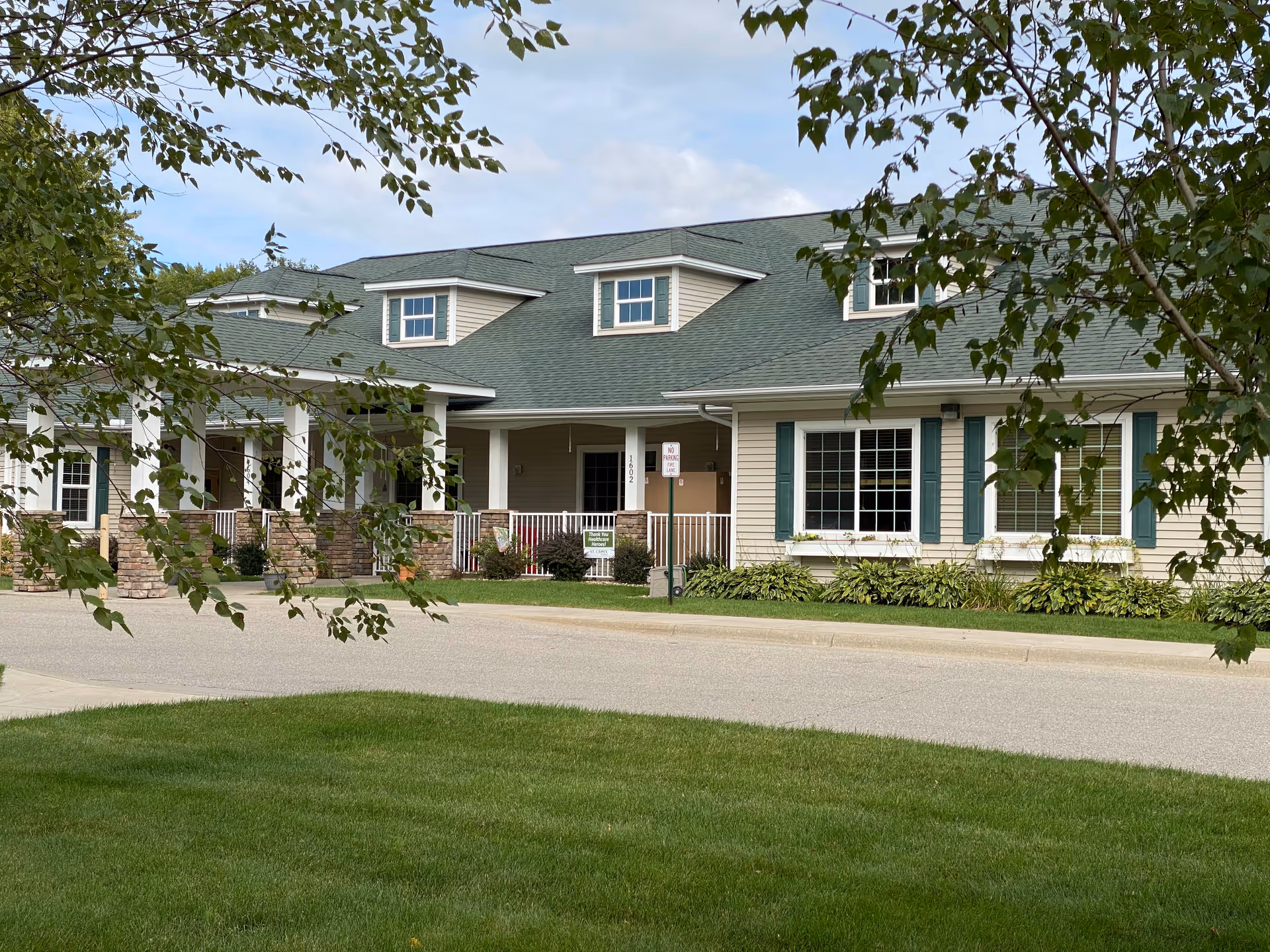 Exterior view of Prairie Senior Cottages of Albert Lea, showing a single-story building with beige siding, green shutters, and a green roof. The building has a covered porch with white columns and stone accents. There is a well-maintained lawn and some trees partially framing the view.