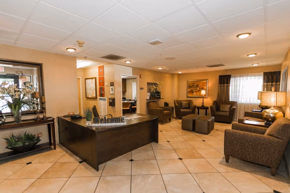 A welcoming senior living facility reception area with a dark wooden desk displaying a 'Welcome' sign, a large mirror on the wall, and a seating area with armchairs, ottomans, and lamps near a window with curtains.