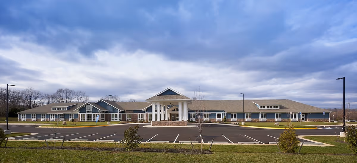 Wide exterior view of a single-story senior living facility building with a large covered entrance, multiple windows, and a spacious parking lot in front under a cloudy sky.