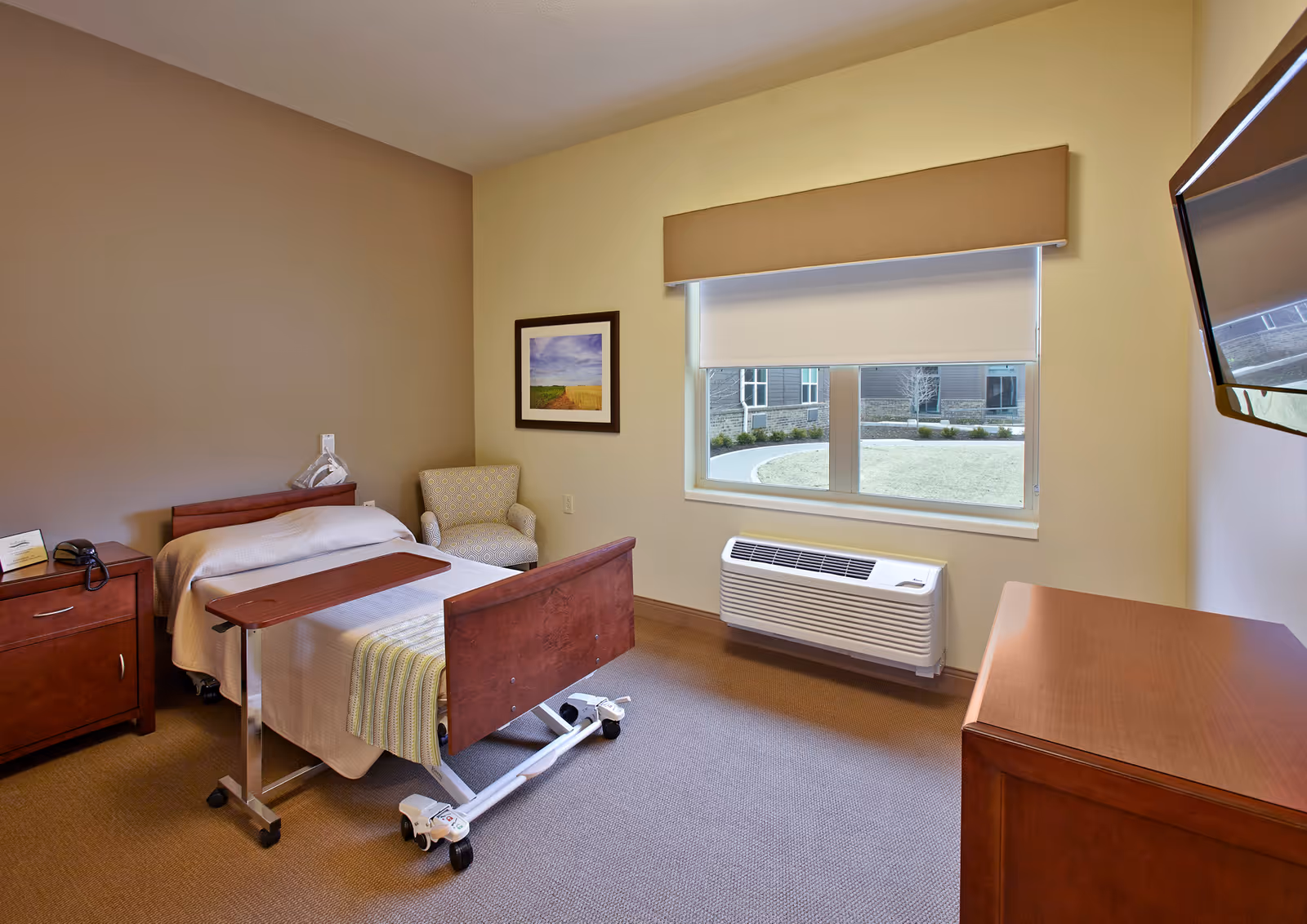 A simple, clean bedroom in a senior living facility with a single hospital-style bed, a wooden nightstand with a phone, a small upholstered armchair, a framed picture on the wall, a large window with a beige valance and a white roller shade, a wall-mounted air conditioning unit below the window, and a flat-screen TV mounted on the wall opposite the bed.