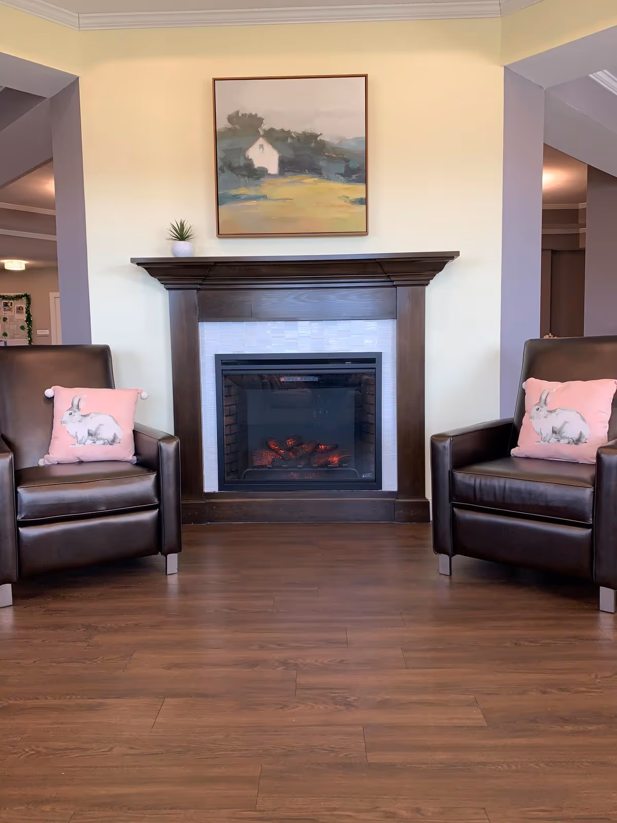 A cozy interior seating area featuring a dark wood fireplace with a white tile surround. Above the fireplace hangs a framed painting of a house in a landscape. On either side of the fireplace are two dark brown leather armchairs, each with a pink pillow decorated with a white rabbit. The floor is a medium-tone wood, and the walls are painted a soft yellow with white crown molding.