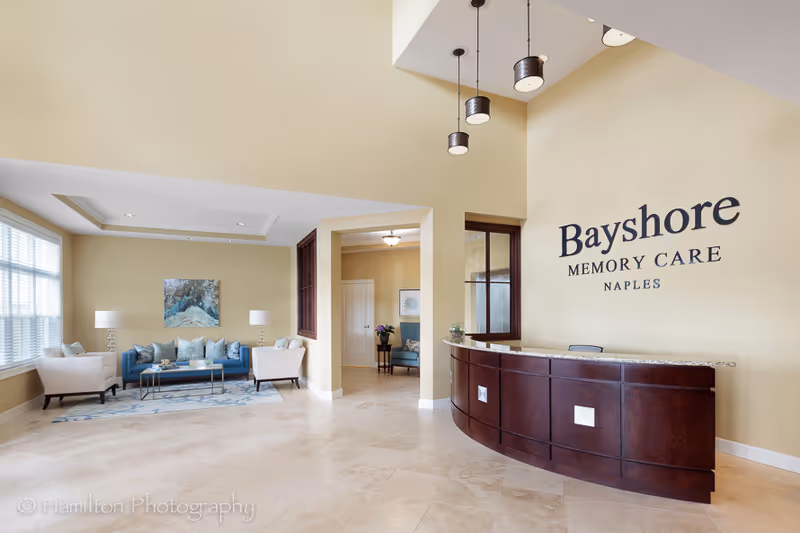 Bright reception lobby with a curved wooden front desk, seating area, and wall sign reading "Bayshore Memory Care Naples".