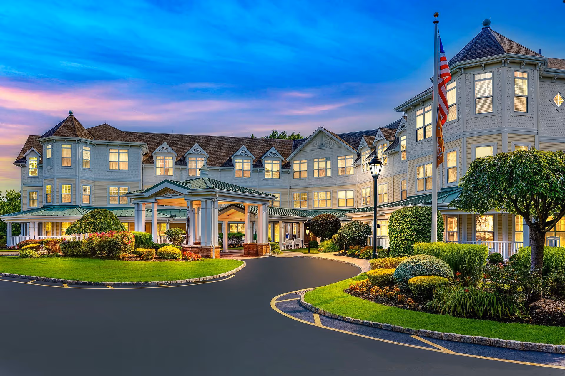 Exterior view of a large, elegant senior living facility building at dusk with well-maintained landscaping, a circular driveway, an American flag, and warm lights glowing from the windows.