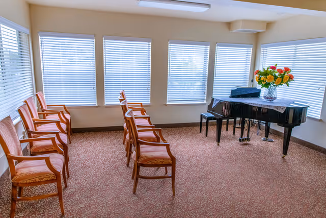 A bright room with several wooden chairs arranged in two rows facing a black grand piano. The room has large windows with white blinds and a vase of colorful flowers on the piano.