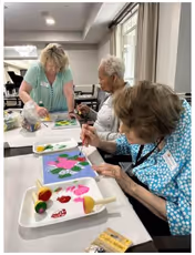 Three elderly women engaged in a painting activity at a table inside a well-lit room. One woman is standing and assisting while the other two are seated, focused on their artwork. Various painting supplies and colorful paint are visible on the table.