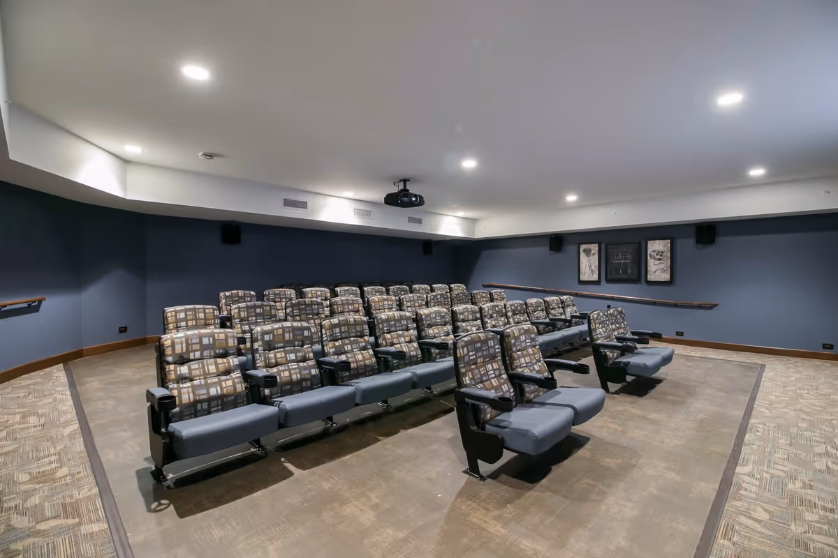 A small theater room with several rows of cushioned seats arranged in a stepped layout facing a projector mounted on the ceiling. The walls are painted dark blue and decorated with framed artwork. The room is well-lit with recessed ceiling lights.