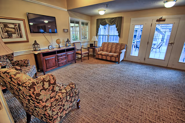 A cozy living room area with patterned armchairs, a small sofa, wooden side tables with lamps, a wooden cabinet with decorative items, a wall-mounted flat screen TV, and large windows and glass doors leading to an outdoor patio.